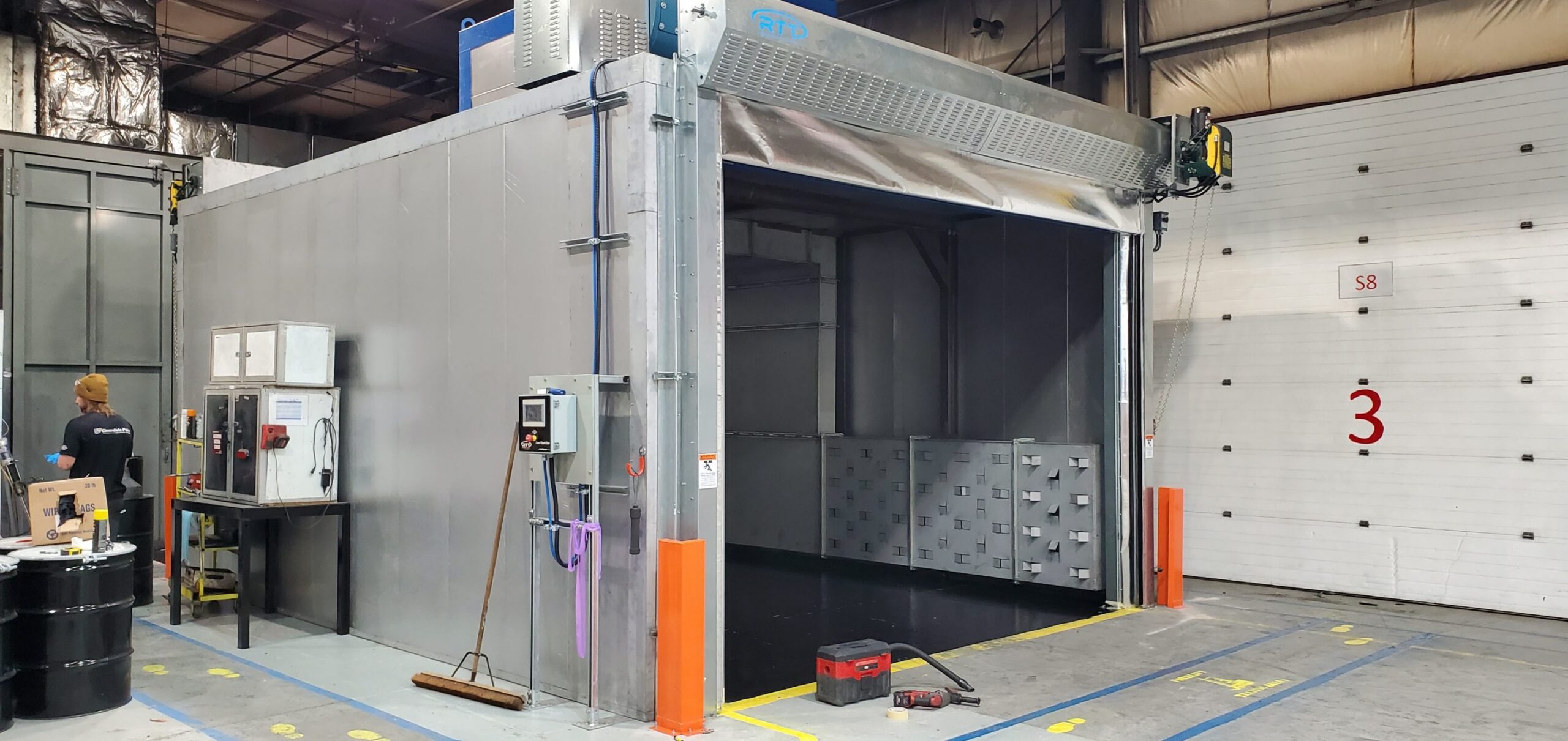 RTT industrial paint booth installed inside a manufacturing facility with large enclosed spray area, overhead ventilation system, control panels, and warehouse workspace visible around the booth.