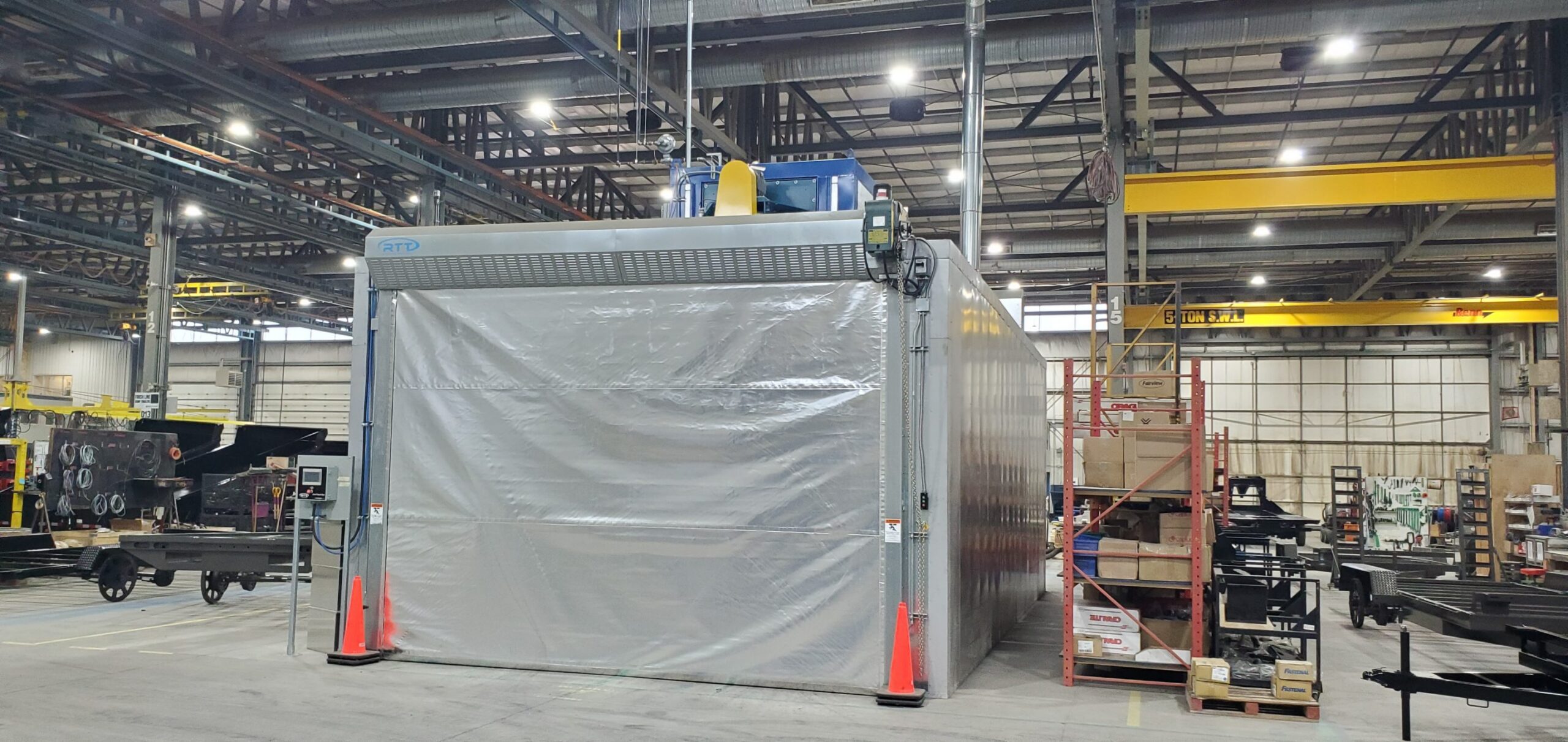 RTT industrial paint booth installed inside a manufacturing facility with large enclosed spray area, overhead ventilation system, control panels, and warehouse workspace visible around the booth.